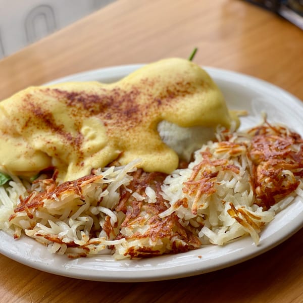 Breakfast Club's Eggs Benedict with hollandaise sauce and hash browns on a white plate.