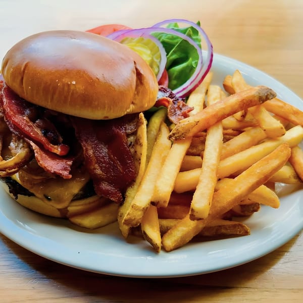 A Breakfast Club bacon cheeseburger with fries, lettuce, tomato, onion, and pickles on a white plate.