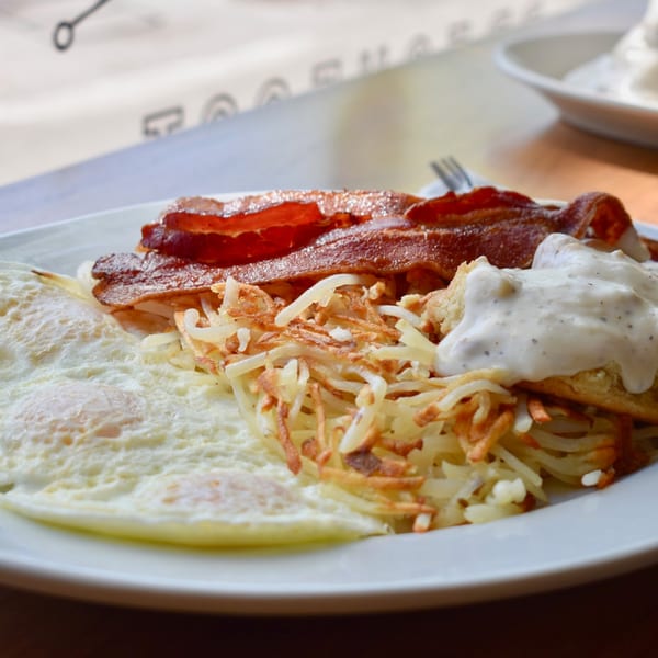 A Breakfast Club plate with crispy bacon, hash browns, fried eggs, and biscuits with gravy.