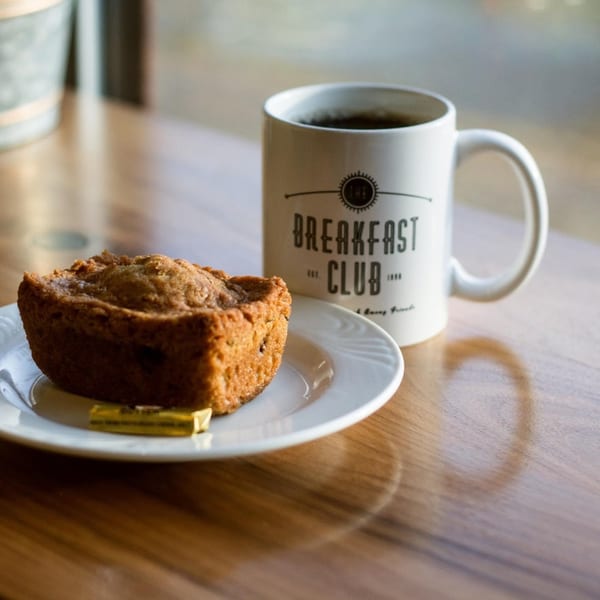 A Breakfast Club coffee mug and a muffin with butter on a white plate on a wooden table.